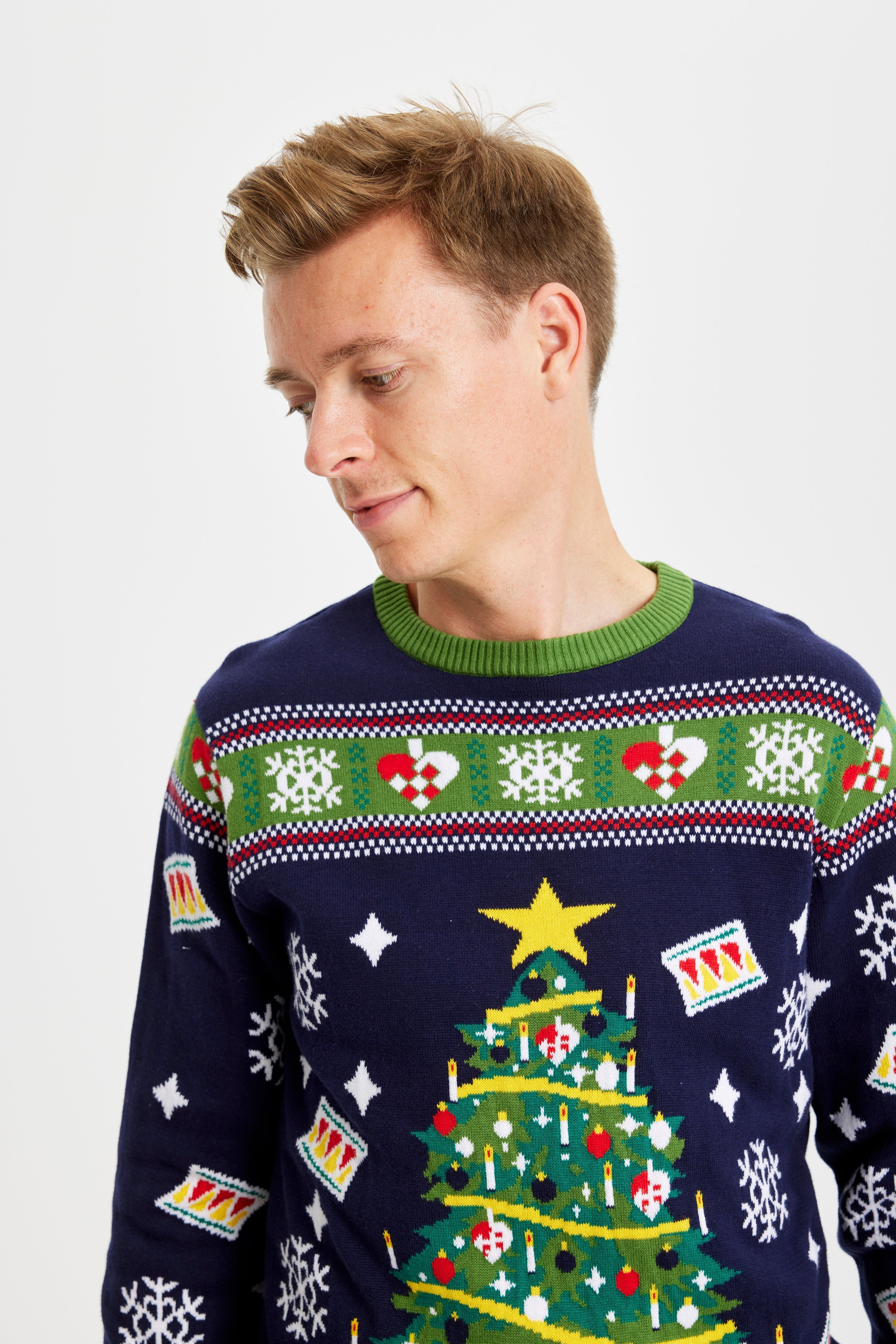 Un homme regarde sur le côté, vêtu d'un pull de Noël bleu et vert orné d'un sapin de Noël.